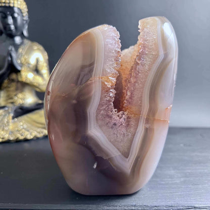 Polierte Achat-Freeform mit zartem Druzy-Innenleben in Rosa und Brauntönen neben Buddha-Figur von Happy Minerals