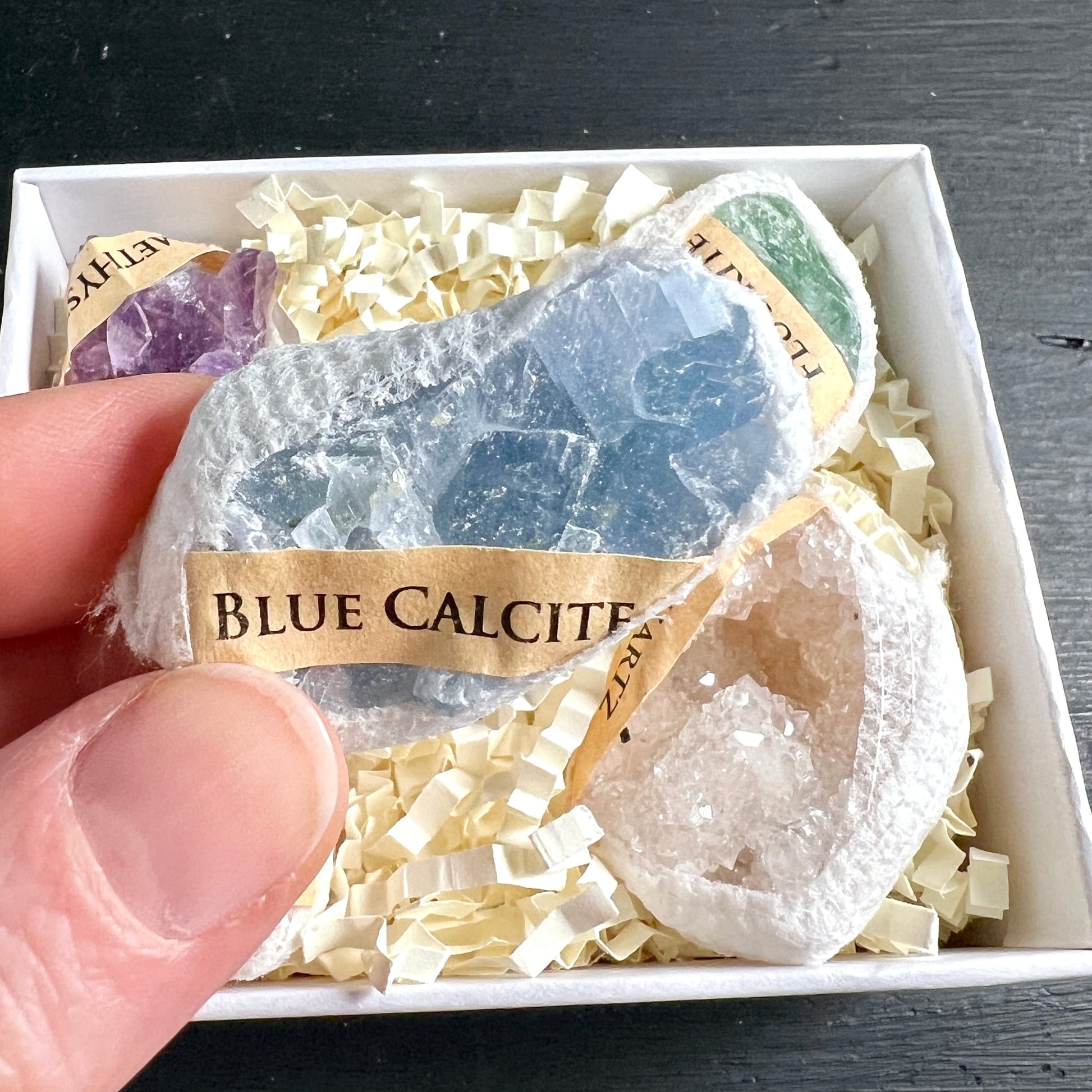 Hand hält Blauer Calcit aus Geschenkbox | lichtblauer Rohstein | Amethyst Fluorit im Hintergrund | Happy Minerals