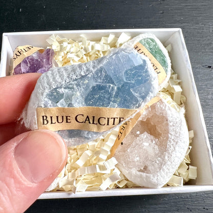 Hand hält Blauer Calcit aus Geschenkbox | lichtblauer Rohstein | Amethyst Fluorit im Hintergrund | Happy Minerals