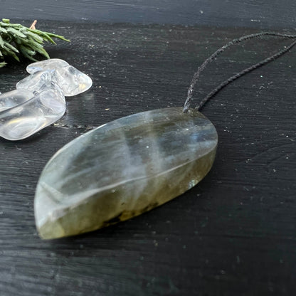 Labradorit Unikat | Rückseite grau-transparent | ovale Blattform | schwarzes Kordel | Kristalle im Hintergrund | Happy Minerals