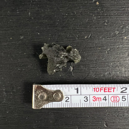 Moldavit Rohstein olivgruen-schwarz mit zerklüfteter Oberflaeche und glasigem Glanz, neben Massband | Happy Minerals