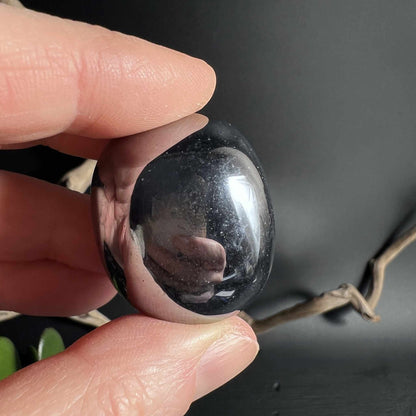 Terahertz Trommelstein ruht zwischen Fingern tiefschwarz mit Lichtspiel | Happy Minerals
